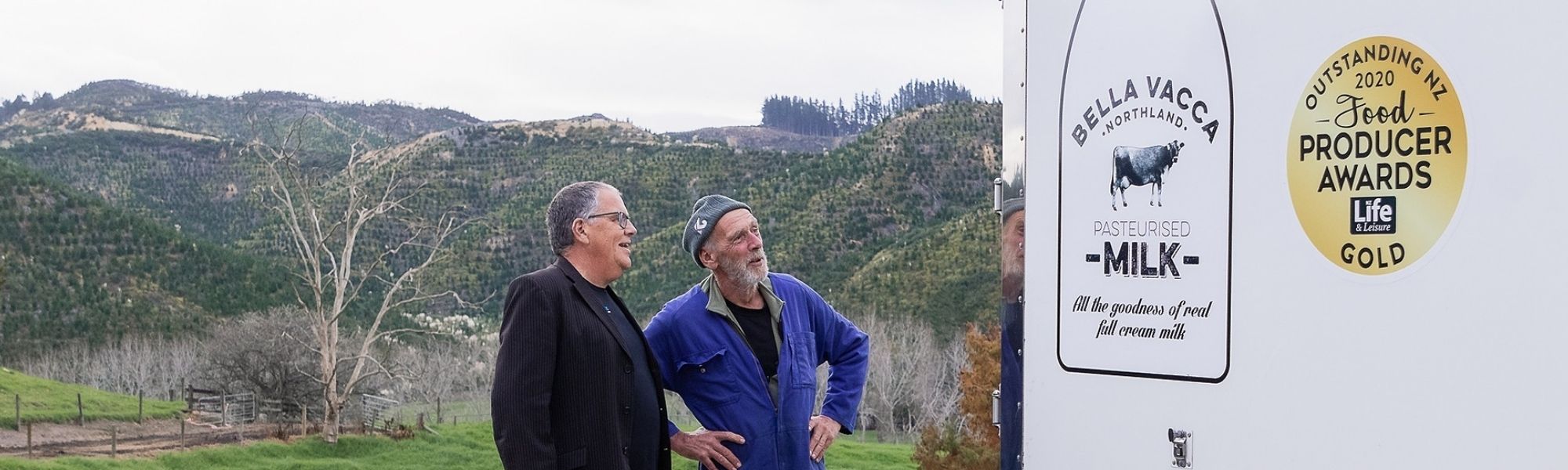 Two men talking in a paddock with tree-planted hills in the background. 